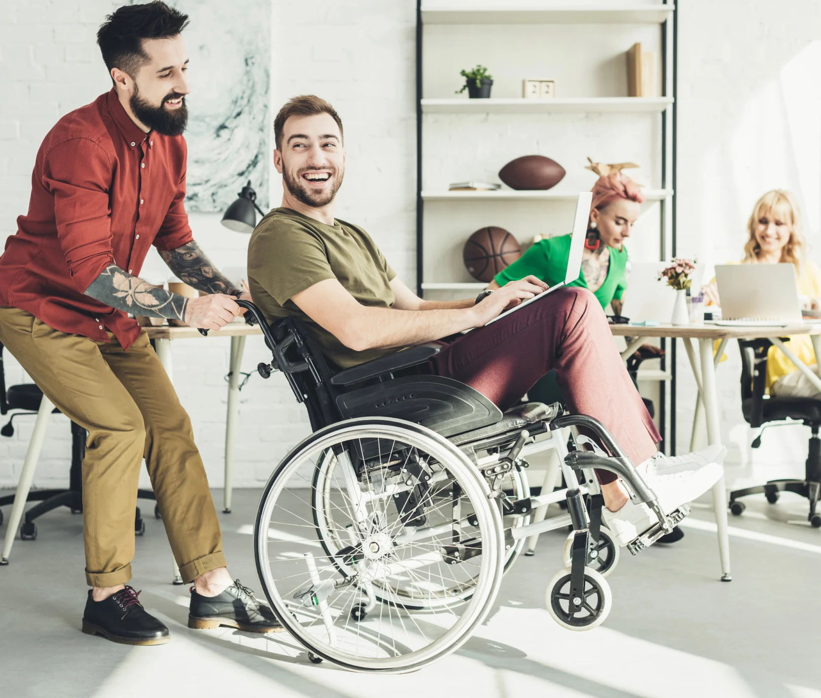 Eine Person im Rollstuhl wird von einem Kollegen durch ein helles Büro geschoben, beide lachen.